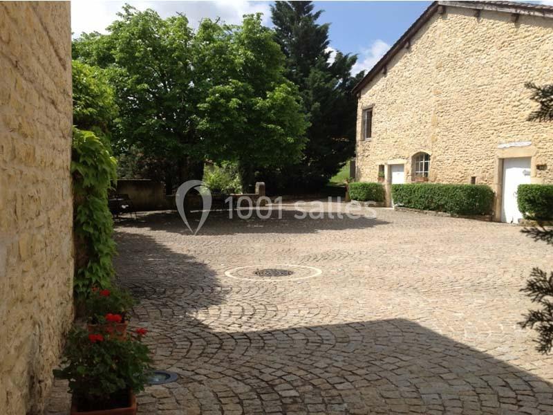 Cour pavée entourée de bâtiments en pierre, avec un arbre au centre et des arbustes en arrière-plan.