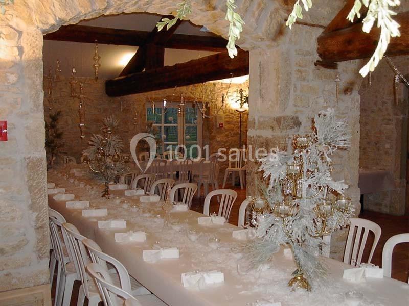 Salle en pierre avec une longue table décorée de centres de table blancs et dorés, entourée de chaises blanches.