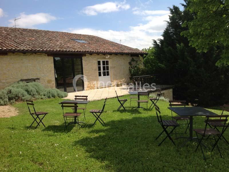 Terrasse en pierre avec tables et chaises en métal sur une pelouse, devant une maison en pierre sous un ciel dégagé.