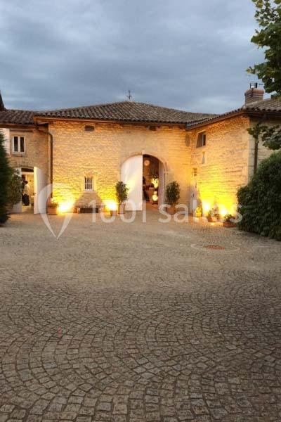 Cour pavée devant un bâtiment en pierre éclairé par des lumières chaudes, avec ciel nuageux en arrière-plan.