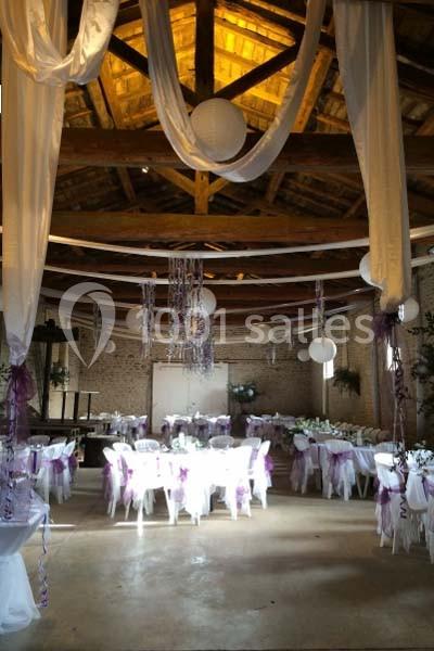Salle de réception décorée avec des tables rondes, chaises blanches ornées de rubans violets et guirlandes suspendues.