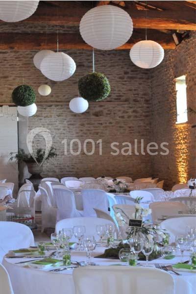 Salle de réception en pierre avec tables dressées, nappes blanches, décorations végétales et lanternes suspendues.