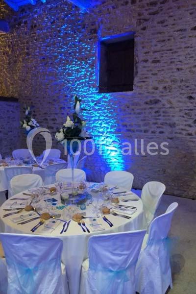 Salle en pierre éclairée de lumières bleues, tables rondes dressées avec nappes blanches et centres de table fleuris.