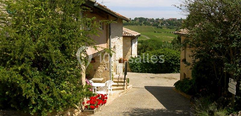 Rue pavée bordée de maisons en pierre avec des fleurs et une vue sur des collines verdoyantes en arrière-plan.