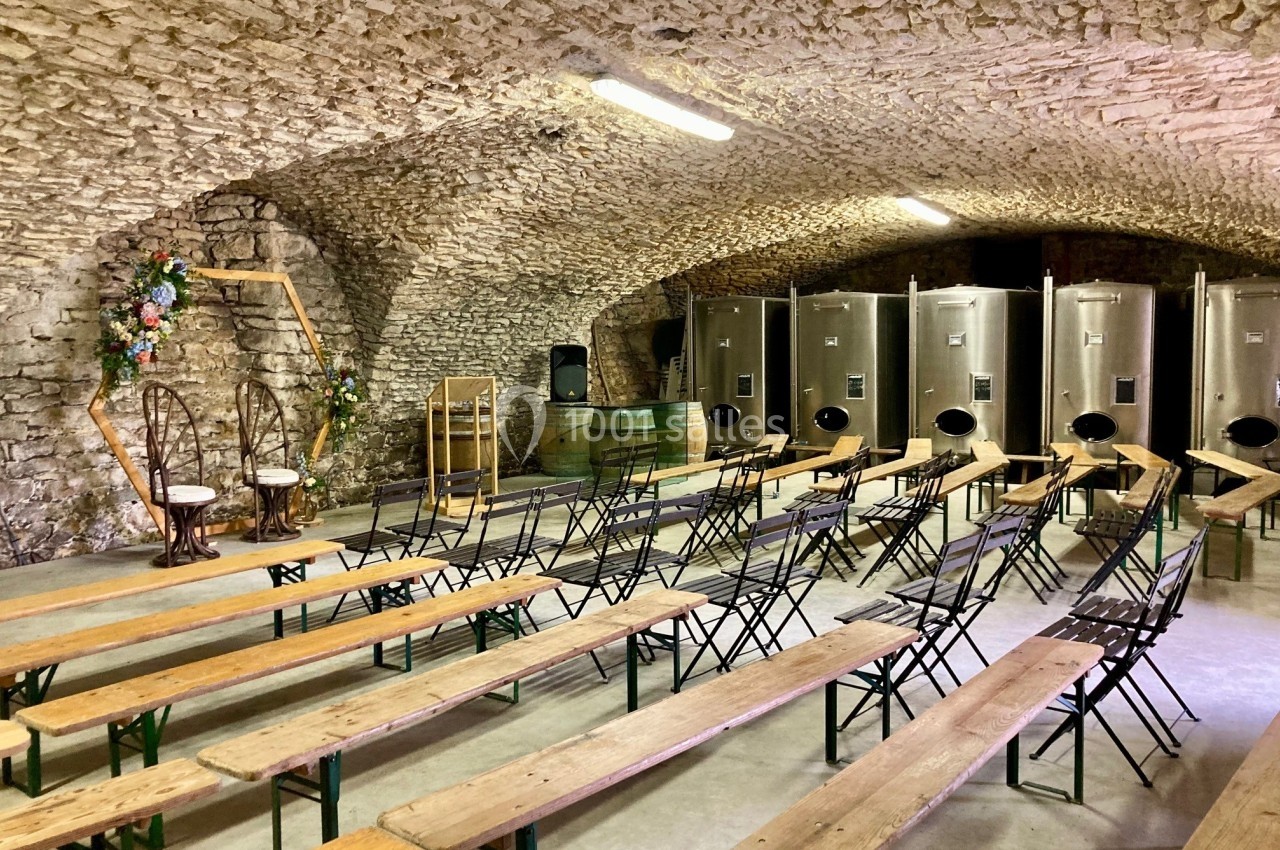 Salle voûtée en pierre avec des bancs en bois, des chaises décorées et des cuves en inox alignées à l'arrière.