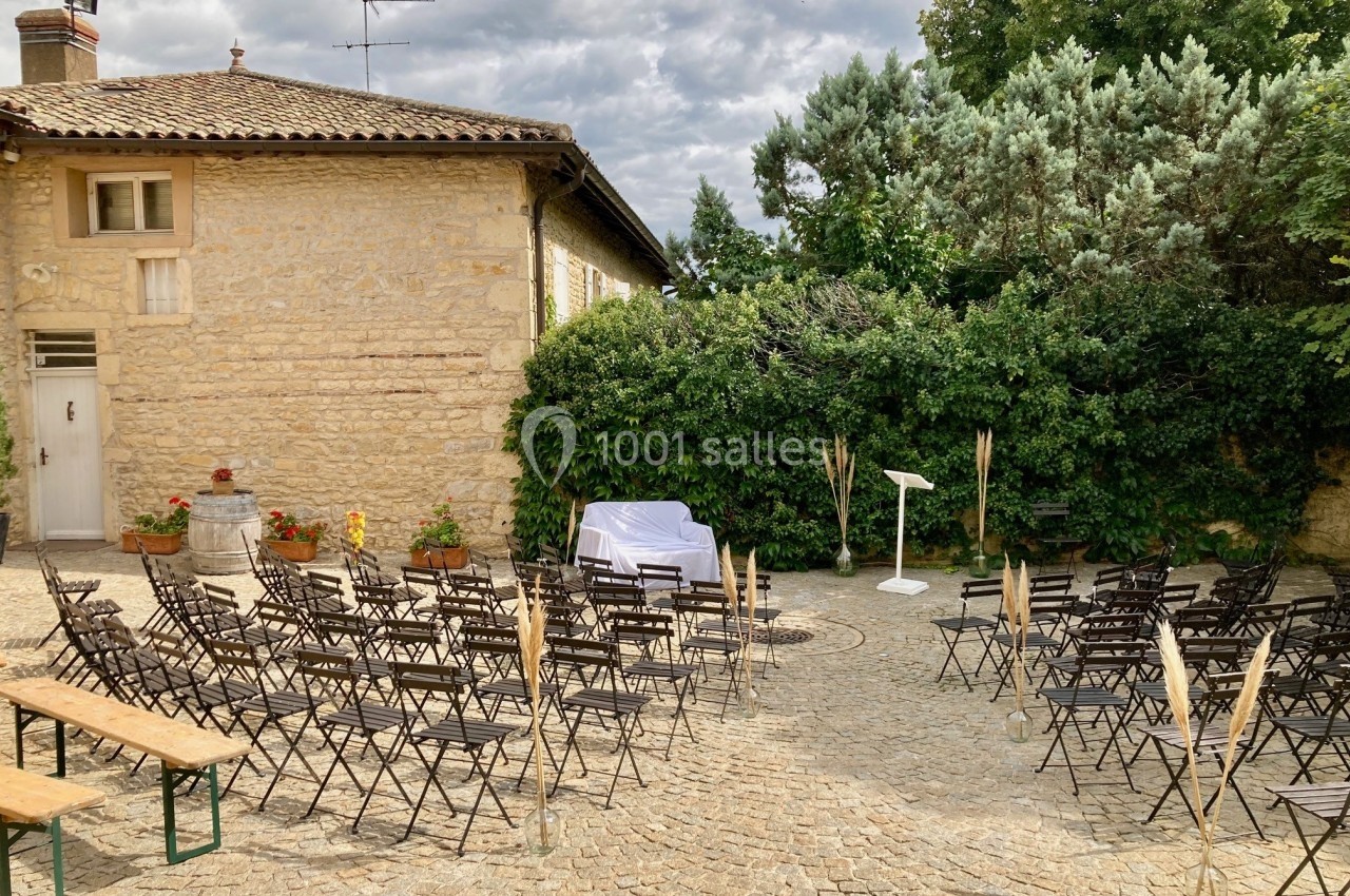 Chaises et bancs disposés en extérieur sur une cour pavée, face à un pupitre blanc, entourés de verdure.
