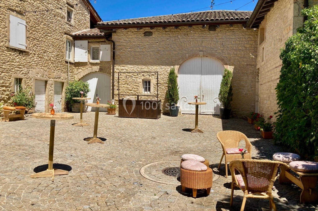 Cour pavée ensoleillée avec tables hautes, chaises en rotin et décorations florales, entourée de bâtiments en pierre.