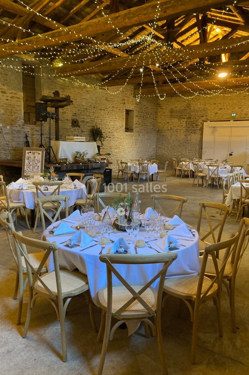 Salle de réception rustique avec tables rondes dressées, chaises en bois et guirlandes lumineuses suspendues au plafond.