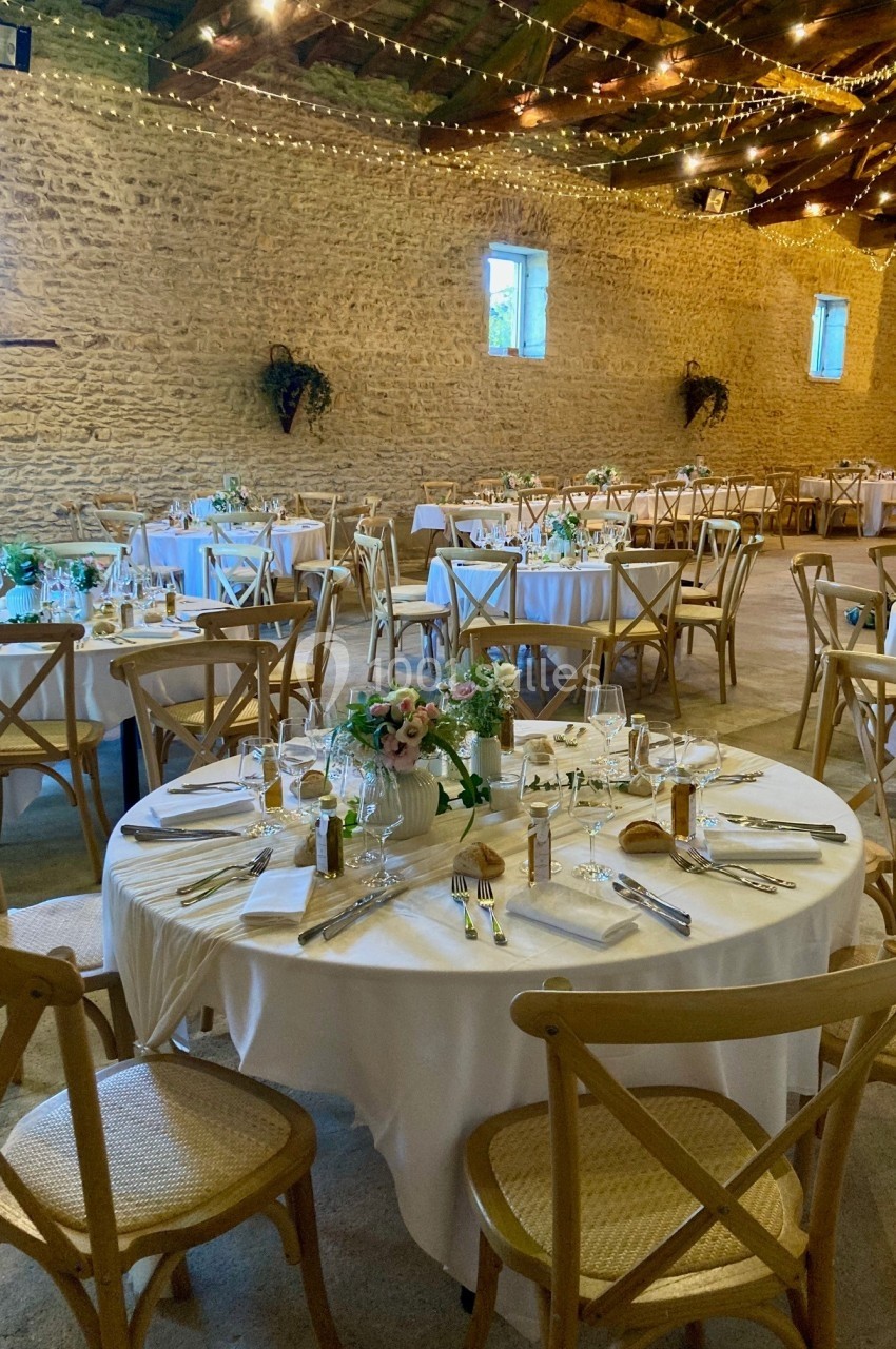 Salle de réception rustique avec tables rondes décorées de nappes blanches, fleurs et couverts, sous des guirlandes…