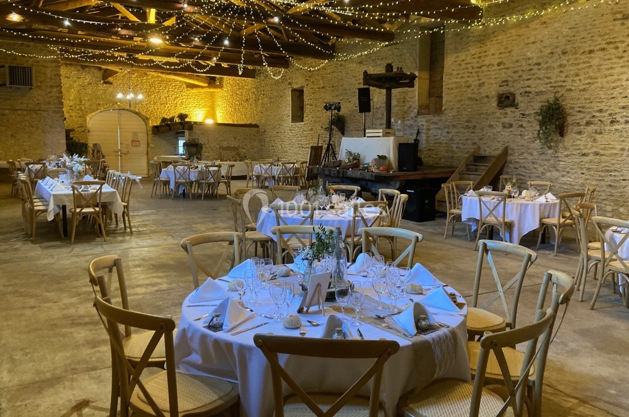Salle de réception rustique avec tables rondes dressées, chaises en bois et guirlandes lumineuses suspendues.