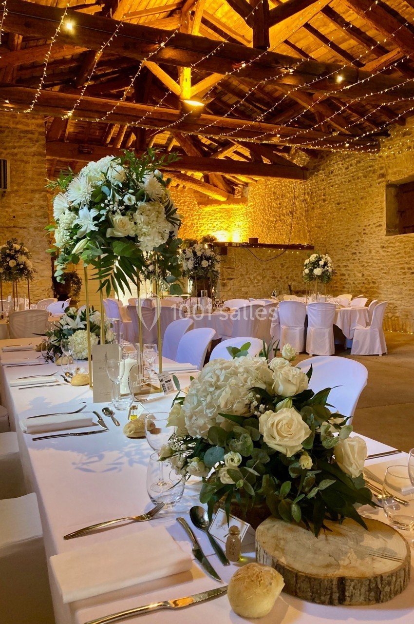 Salle de réception en pierre avec tables décorées de fleurs blanches, nappes blanches et éclairage chaleureux.