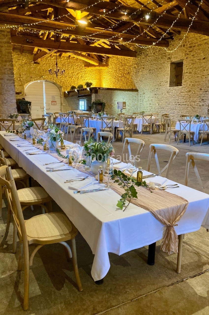 Salle de réception rustique avec tables décorées de nappes blanches, plantes et lumières suspendues au plafond en bois.