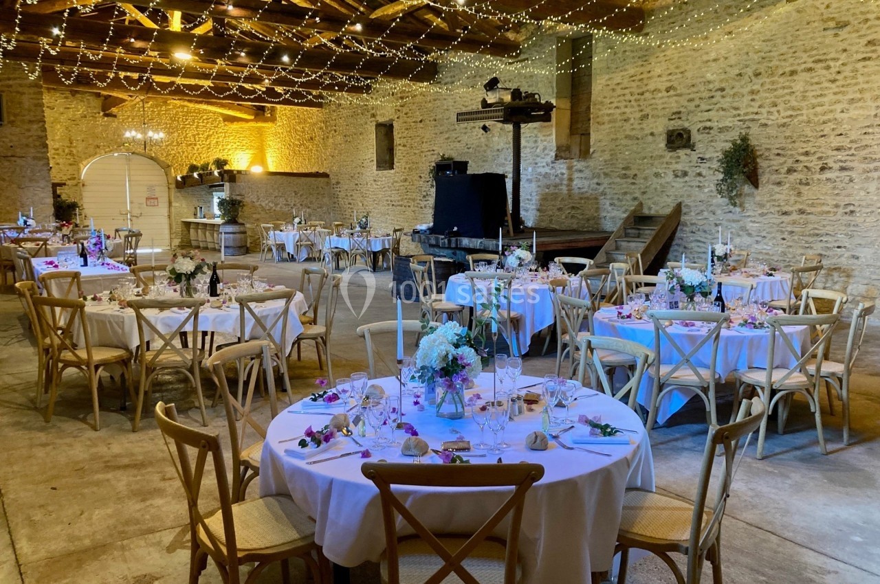 Salle de réception rustique décorée avec des guirlandes lumineuses, des tables rondes dressées et des chaises en bois.