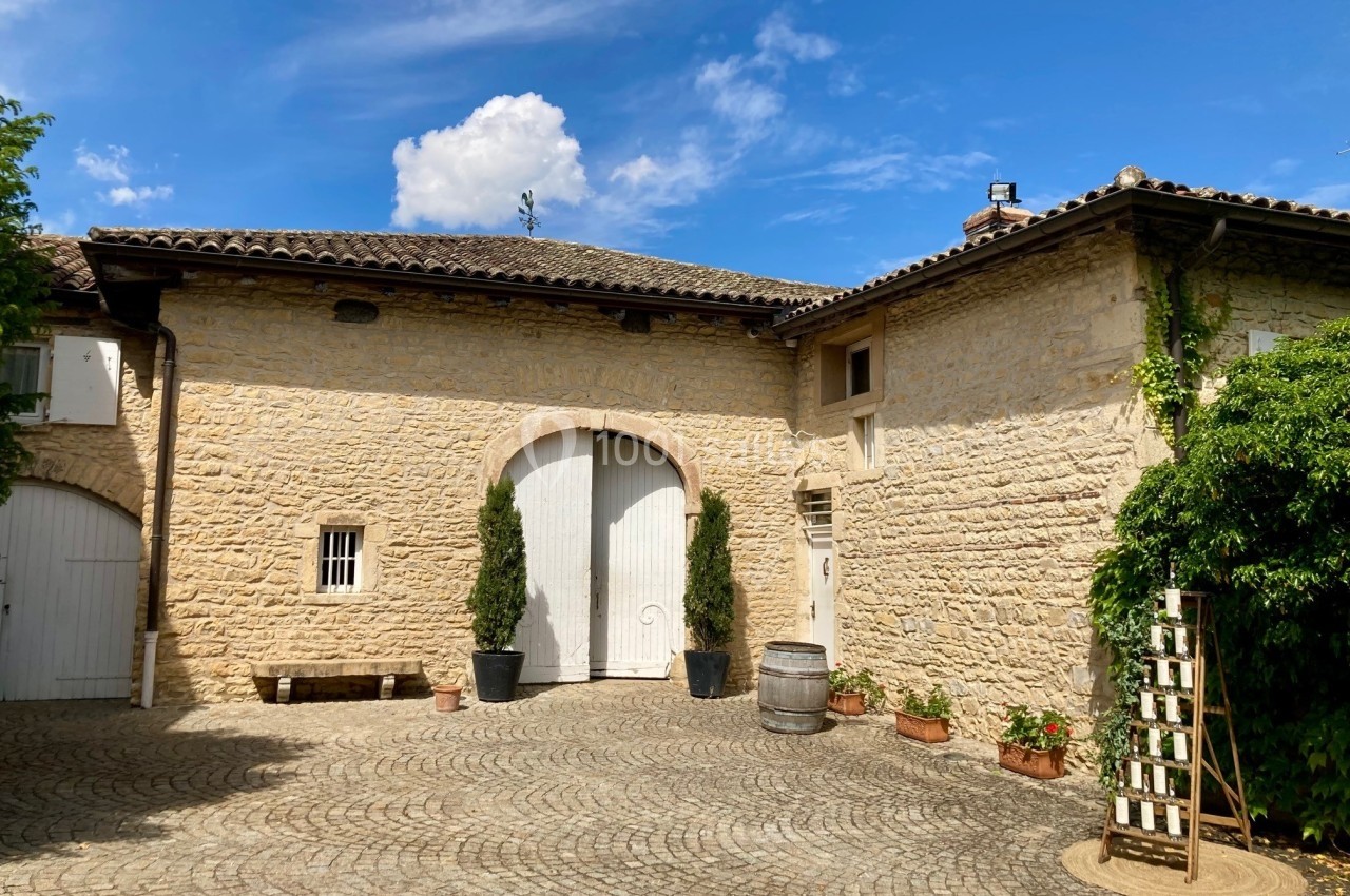 Cour pavée avec façade en pierre, portes blanches cintrées, pots de plantes et ciel bleu en arrière-plan.