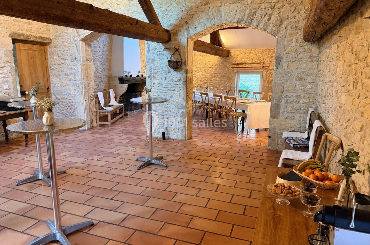 Salle en pierre avec poutres apparentes, tables dressées, chaises en bois et buffet avec fruits et boissons.