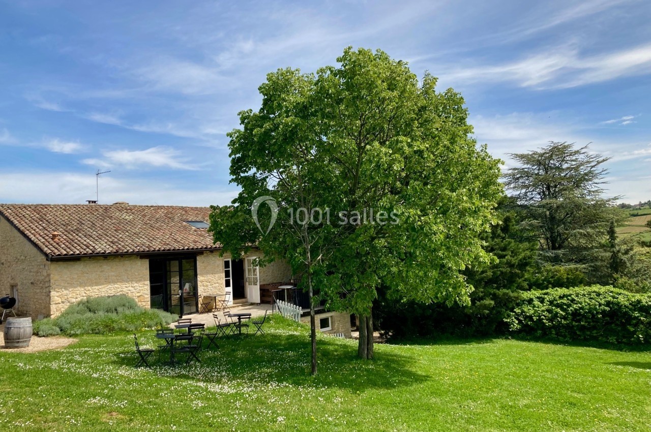 Maison en pierre avec toit en tuiles, entourée d'un jardin verdoyant, un arbre au premier plan et ciel dégagé.