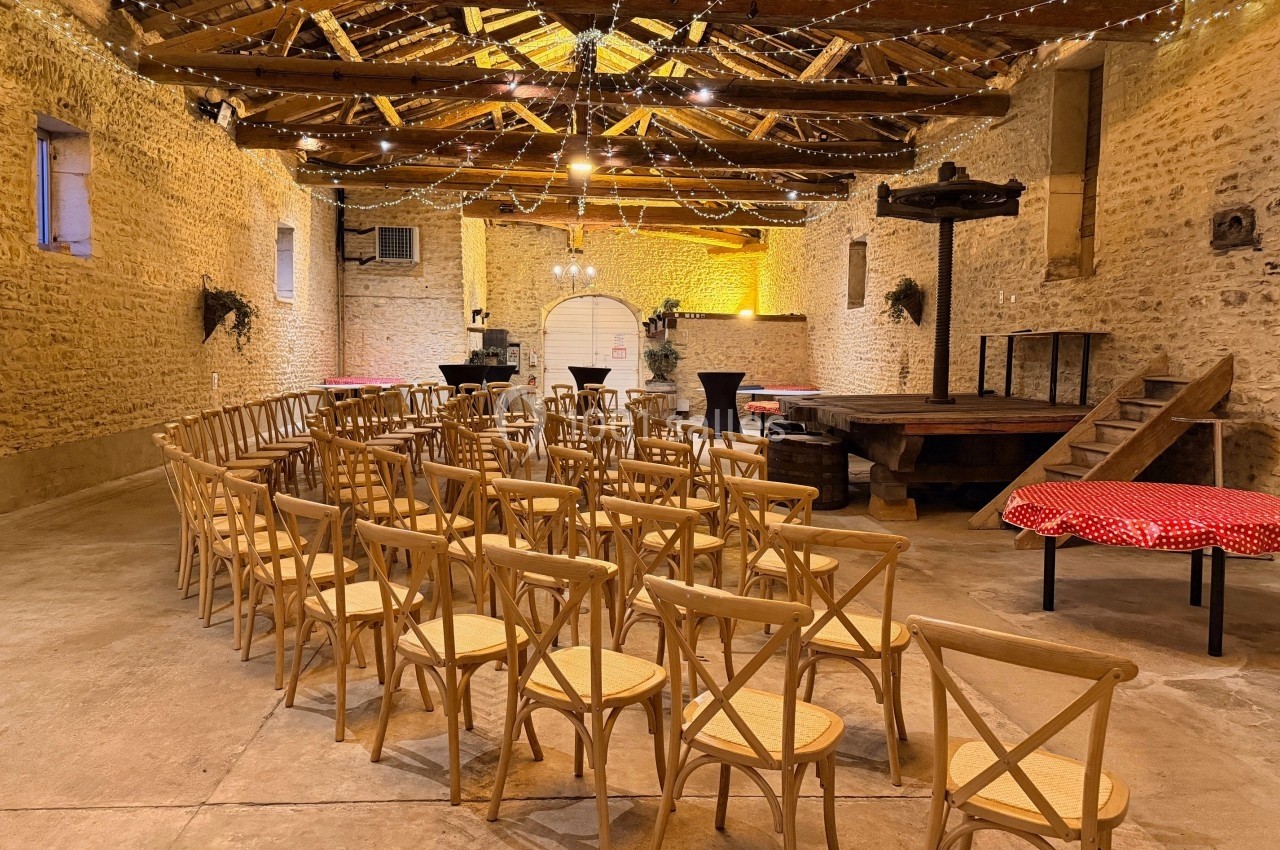 Salle en pierre avec des chaises en bois disposées face à une petite scène, éclairée par des guirlandes lumineuses.