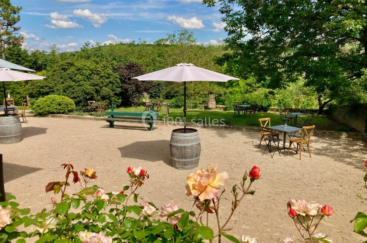 Terrasse extérieure avec tables, chaises et parasols, entourée de verdure et de fleurs sous un ciel ensoleillé.
