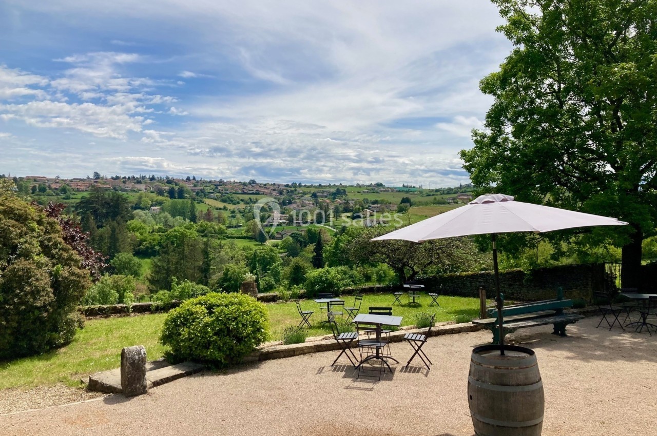 Terrasse avec tables et parasol, offrant une vue dégagée sur un paysage vallonné et verdoyant sous un ciel partiellement…