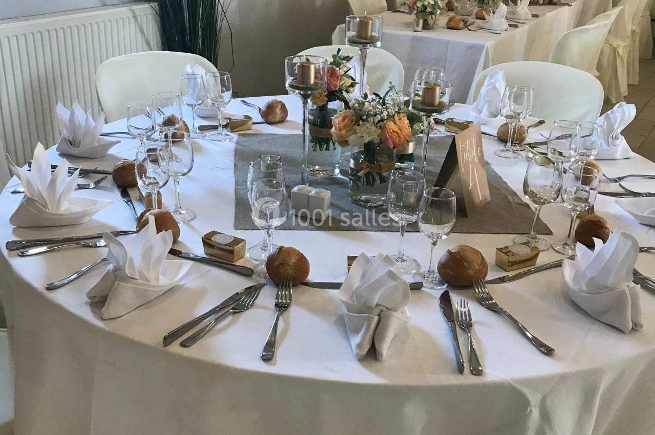 Table ronde dressée pour un événement, avec nappes blanches, serviettes pliées, vaisselle, bougies et décoration florale.