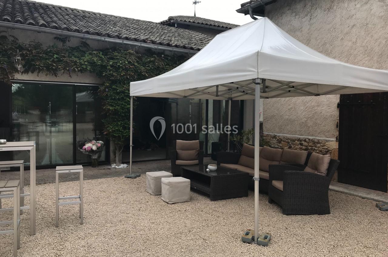 Tonnelle blanche installée dans une cour en gravier, avec salon de jardin en rotin et murs en pierre en arrière-plan.