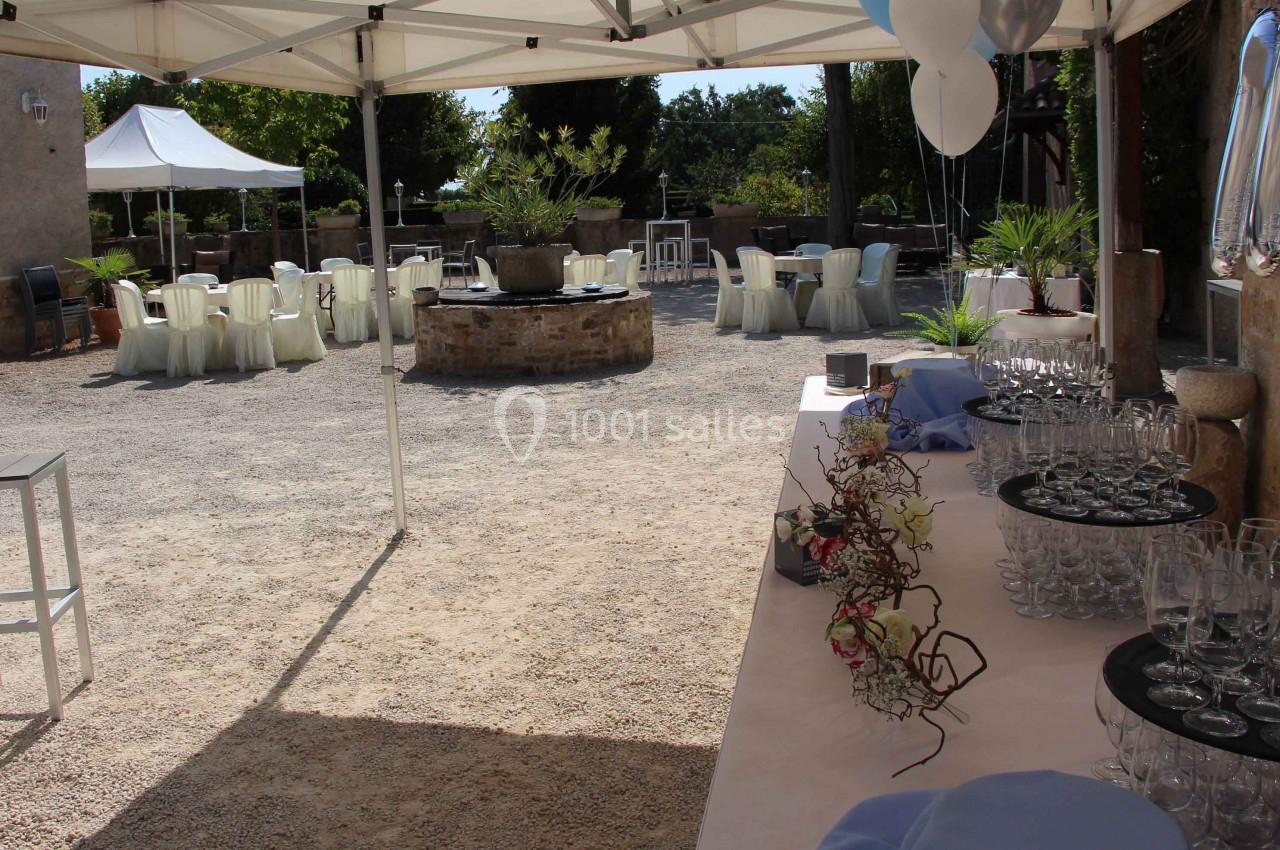 Table dressée avec verres et décorations florales sous un chapiteau, avec des tables rondes disposées en extérieur.