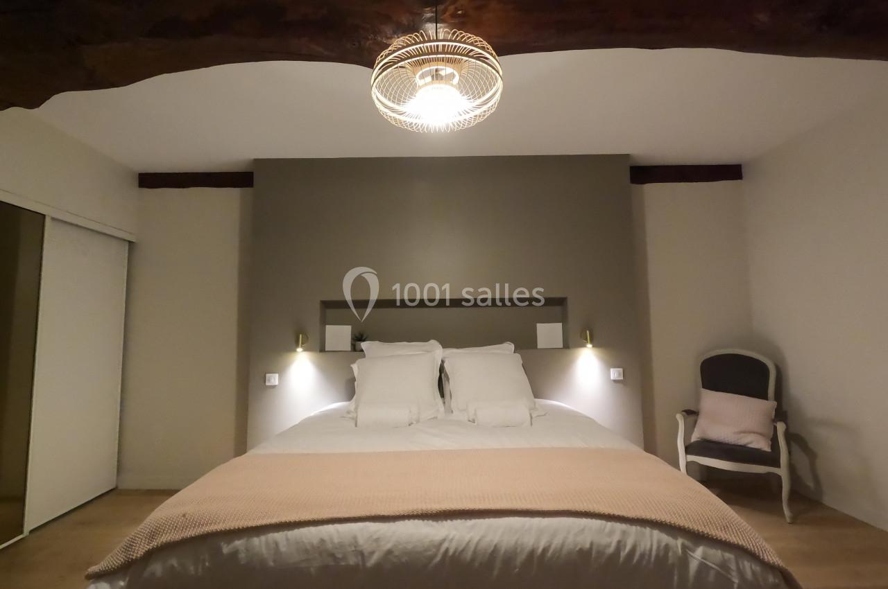 Lit double avec couverture beige dans une chambre lumineuse, éclairée par une suspension et décorée de poutres apparentes.