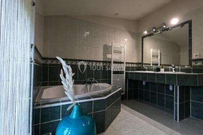 Petit pont en fer forgé traversant un cours d'eau bordé d'arbres, près d'une maison en pierre avec toit en ardoise.