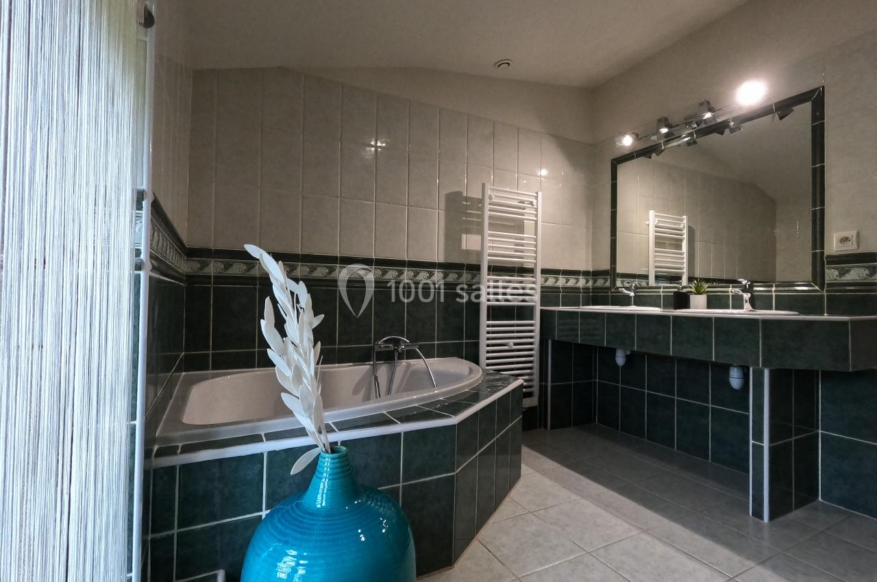 Salle de bain avec baignoire d'angle, carrelage vert et blanc, miroir éclairé et vase décoratif turquoise.