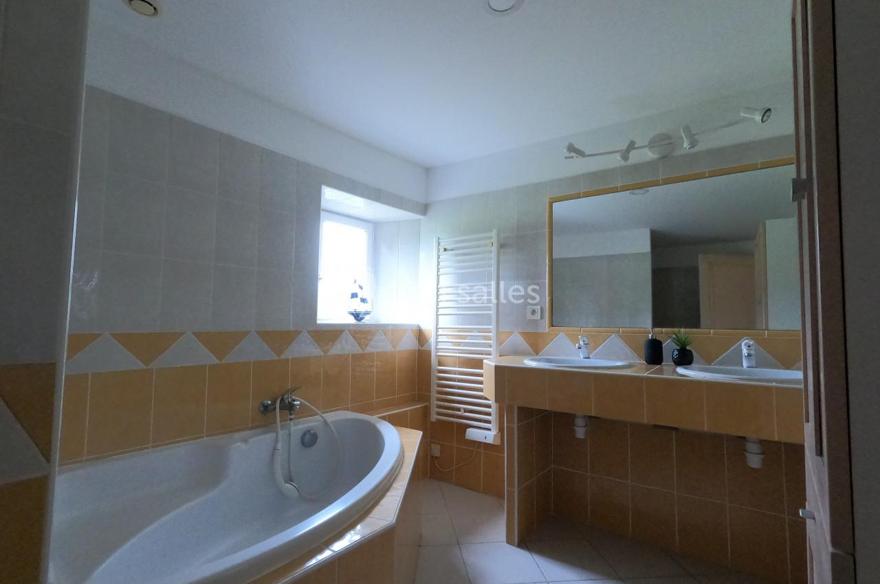 Salle de bain lumineuse avec baignoire d'angle, double vasque, miroir mural et carrelage beige et blanc.