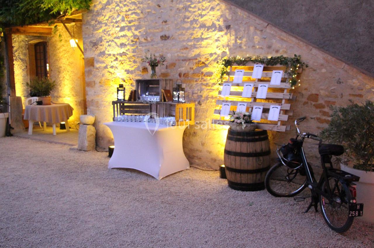 Décoration de mariage en extérieur avec table de boissons, plan de table sur tonneau et vélo appuyé contre un mur en pierre.