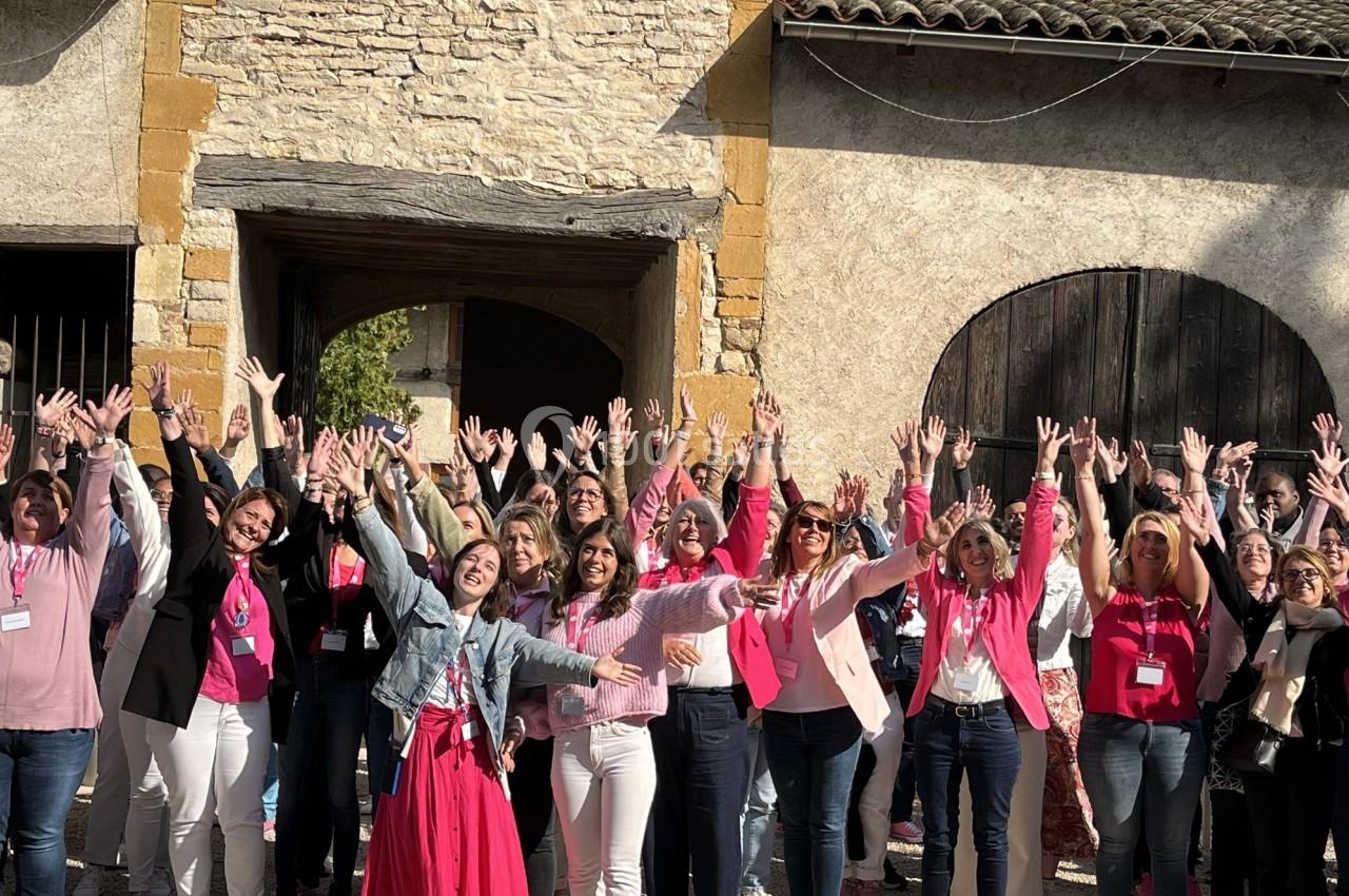 Un groupe de personnes souriantes levant les bras devant des bâtiments en pierre sous un ciel ensoleillé.