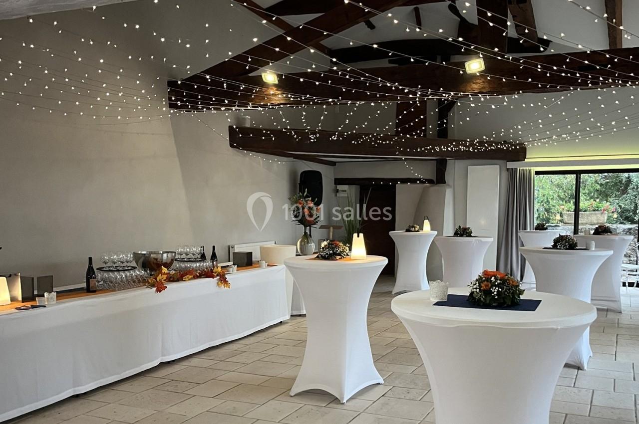 Salle décorée pour un événement avec guirlandes lumineuses, tables hautes nappées de blanc et buffet dressé.
