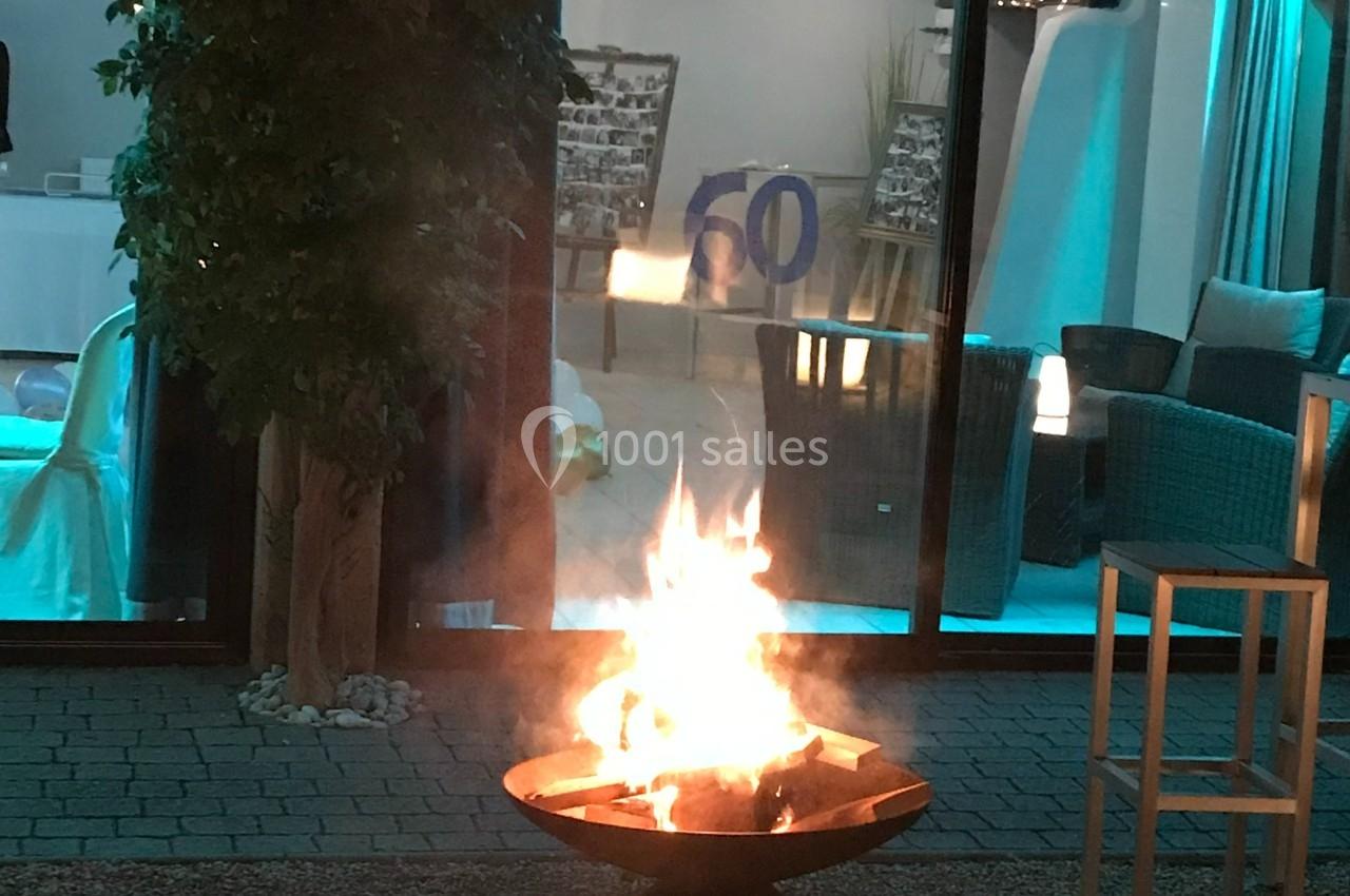 Brasero allumé sur une terrasse en soirée, devant une baie vitrée éclairée et un décor intérieur.