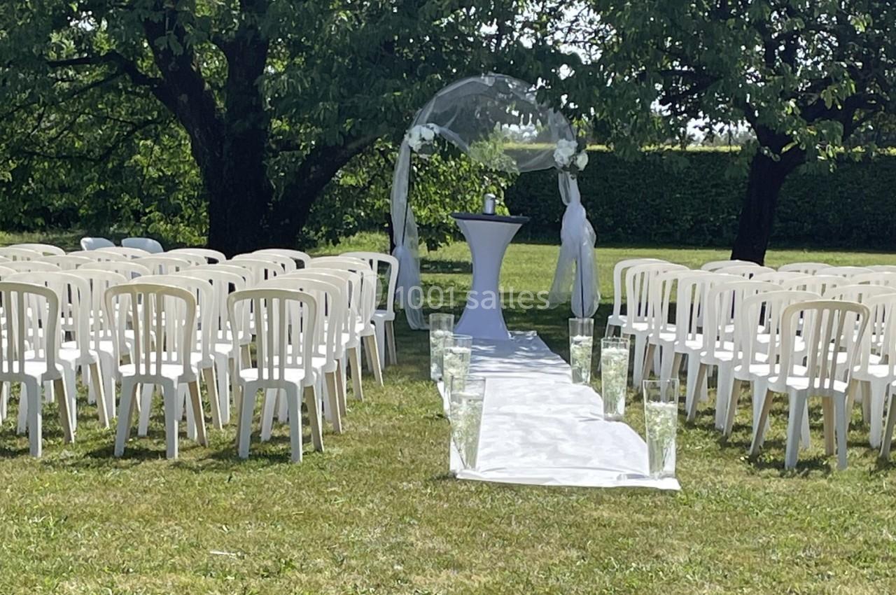 Allée centrale décorée pour une cérémonie en plein air, entourée de chaises blanches et d'arbres.