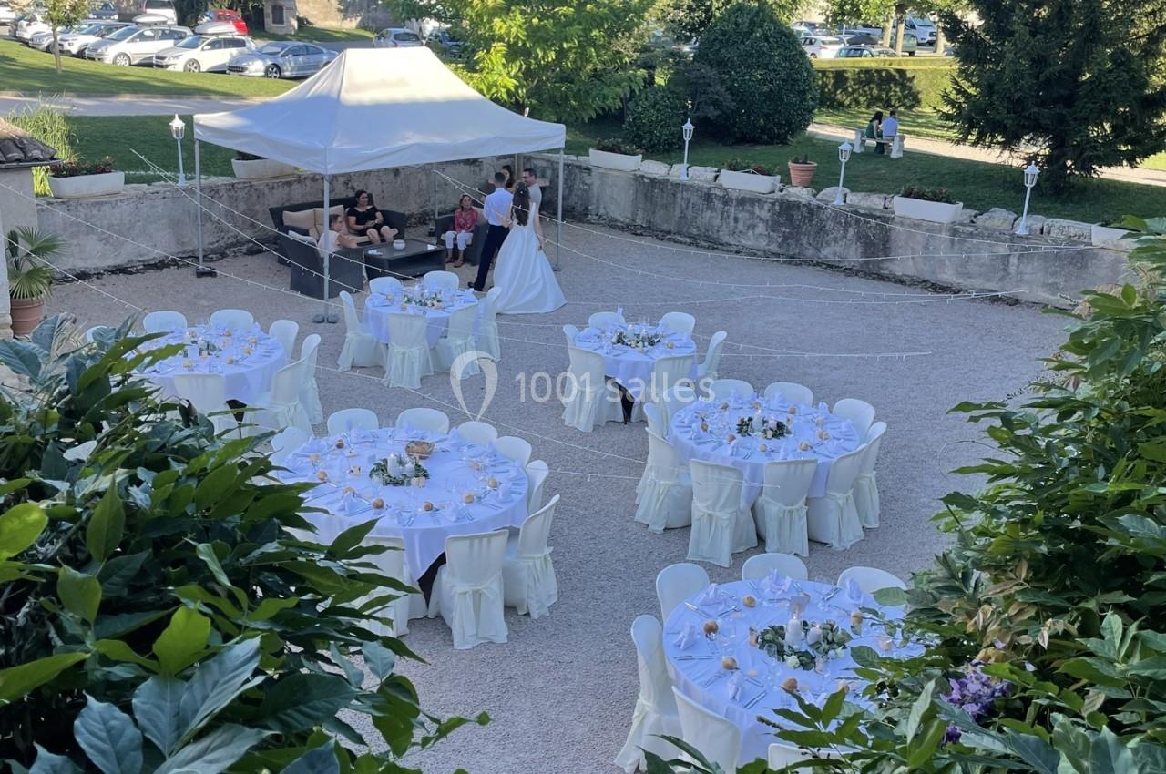 Vue d'une réception en plein air avec tables rondes dressées et un couple en tenue de mariage près d'une tente.