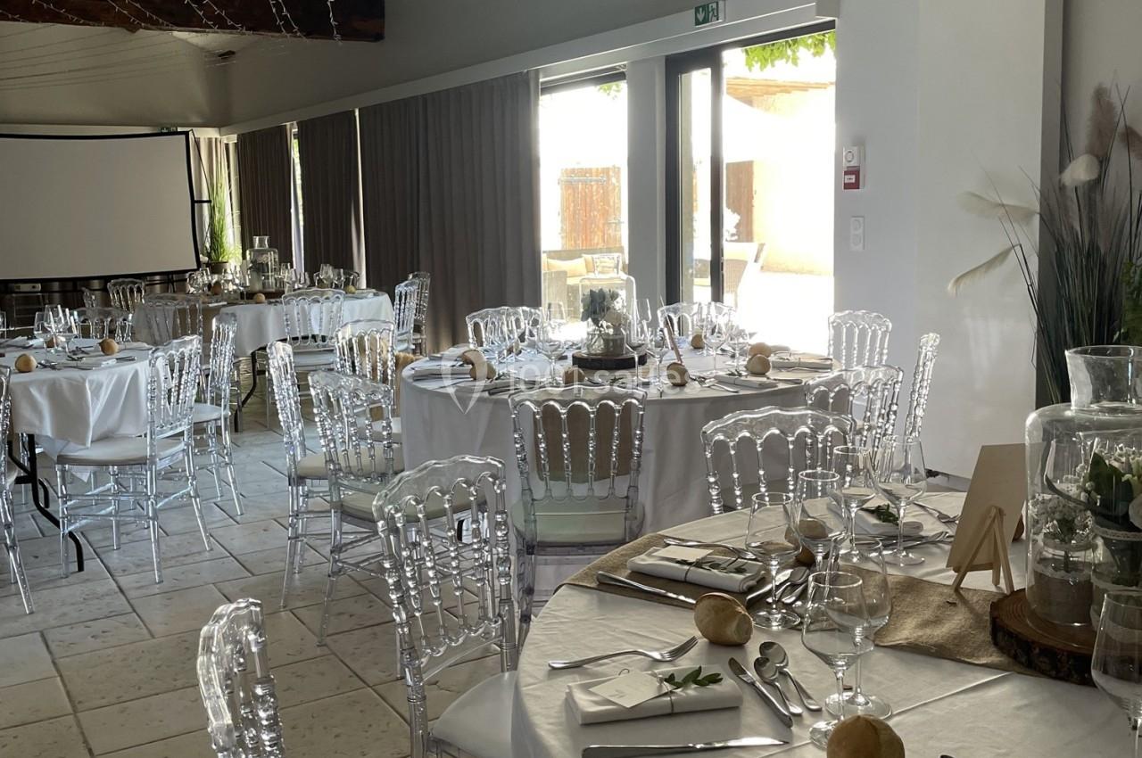 Salle de réception décorée avec des tables rondes dressées, chaises transparentes et lumière naturelle entrant par de…