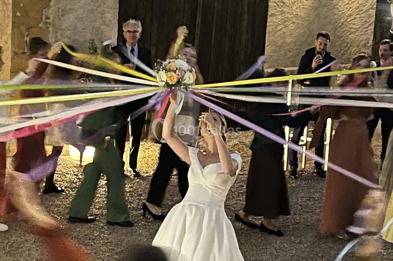 Une mariée en robe blanche tient un bouquet entouré de rubans colorés, avec des invités en arrière-plan.