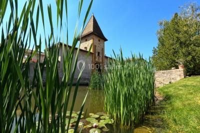 Petit pont en fer forgé traversant un cours d'eau bordé d'arbres, près d'une maison en pierre avec toit en ardoise.