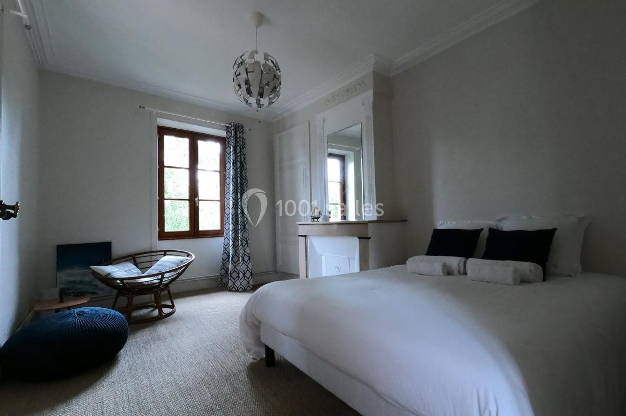 Chambre lumineuse avec lit double, fauteuil en osier, grande fenêtre et cheminée blanche dans un décor épuré.