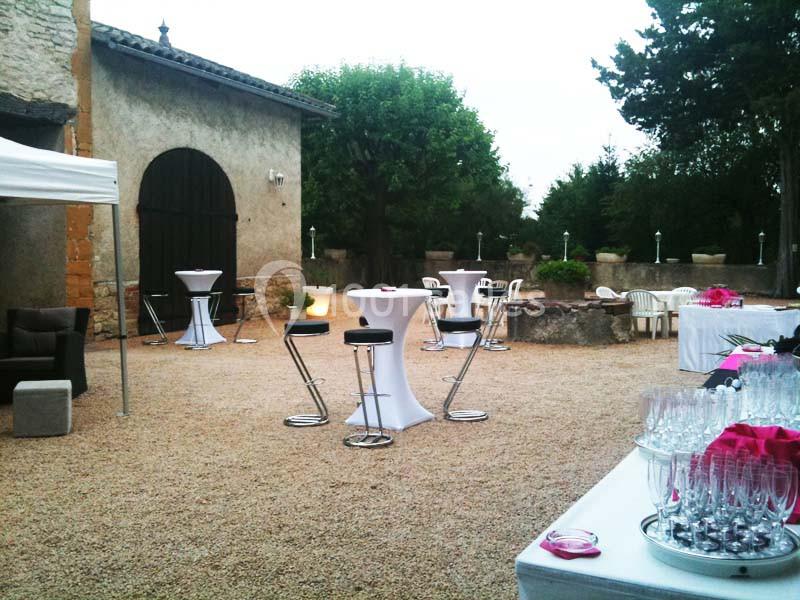 Cour extérieure aménagée pour un événement avec tables hautes, chaises, nappes blanches et décorations roses.