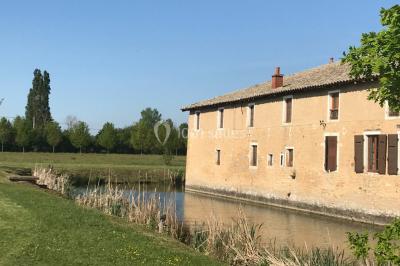 Petit pont en fer forgé traversant un cours d'eau bordé d'arbres, près d'une maison en pierre avec toit en ardoise.