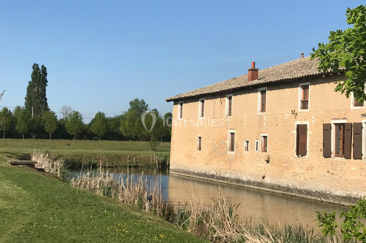 Bâtiment en pierre beige au bord d'un canal, entouré de végétation et sous un ciel bleu clair.
