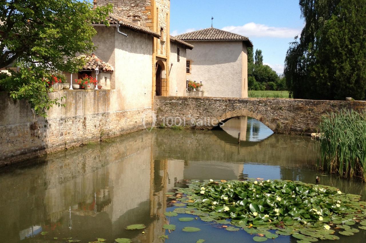 Vue d'un bâtiment ancien avec une tour, entouré d'un fossé d'eau calme, des nénuphars et un petit pont en pierre.