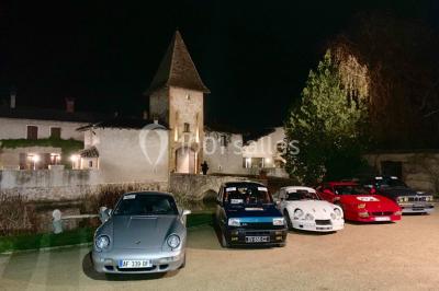 Petit pont en fer forgé traversant un cours d'eau bordé d'arbres, près d'une maison en pierre avec toit en ardoise.