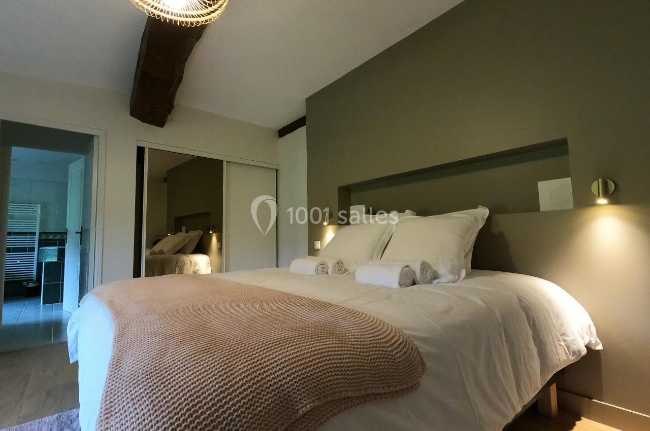 Lit double avec couverture beige, draps blancs et serviettes pliées, dans une chambre lumineuse avec poutre apparente.