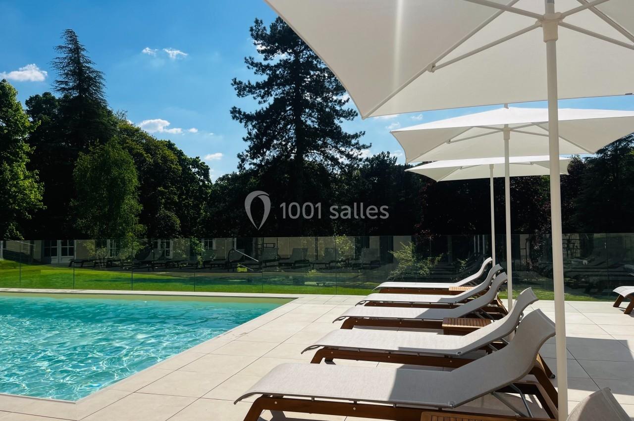 Chaises longues alignées sous des parasols blancs au bord d'une piscine, entourée de verdure et d'arbres.