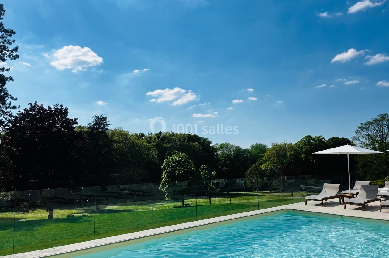 Piscine extérieure avec transats et parasol, entourée d'une pelouse et d'arbres sous un ciel bleu dégagé.