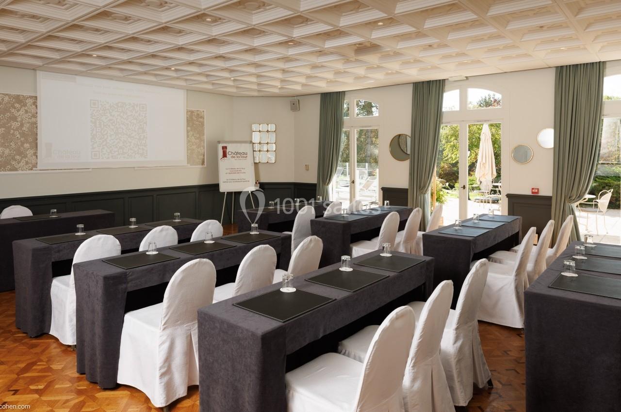 Salle de réunion lumineuse avec des tables alignées, chaises blanches et écran de projection à l'avant.
