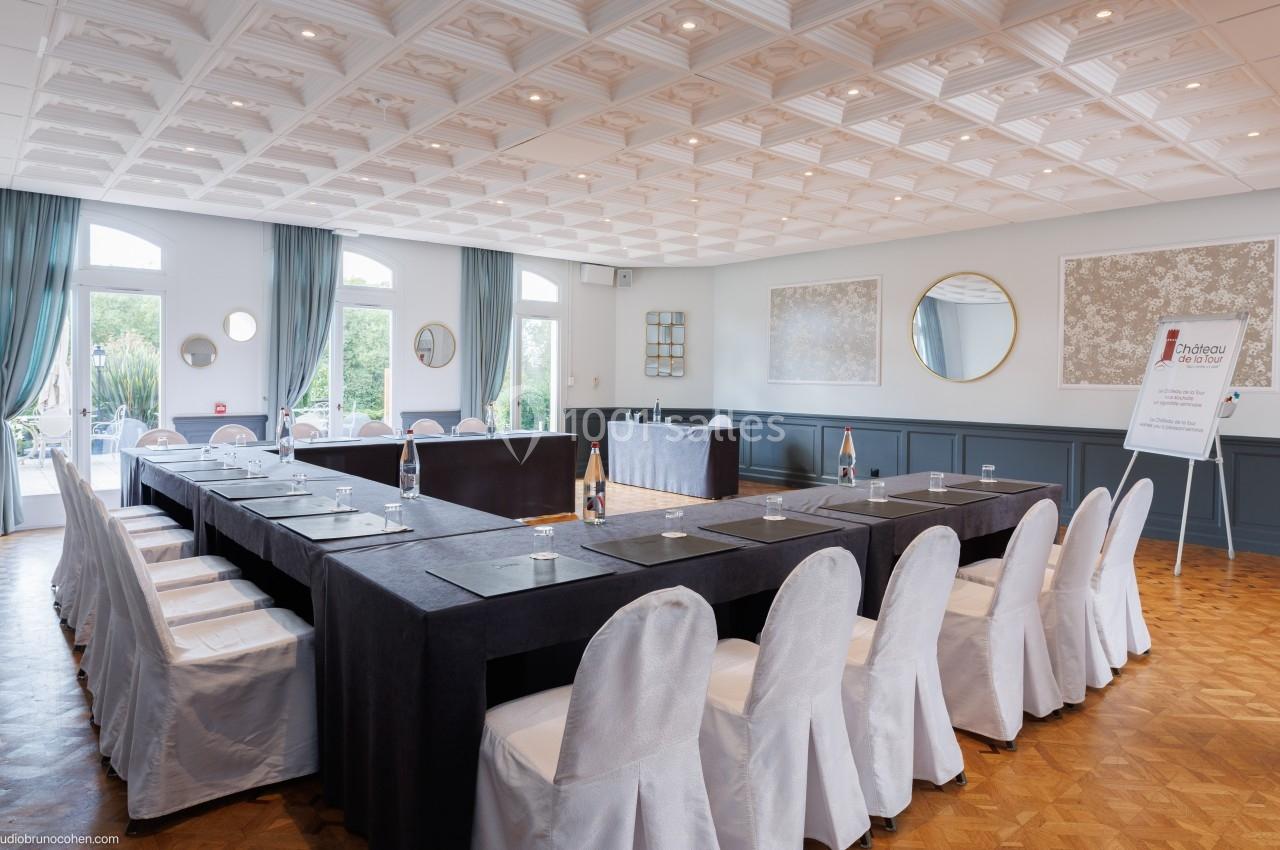 Salle de réunion lumineuse avec tables disposées en U, chaises blanches et grandes fenêtres donnant sur l'extérieur.